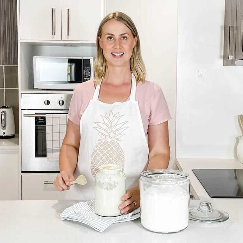 Picture of Emily set in the kitchen with a jar of sourdough starter, a wooden mixing spoon and a container of flour