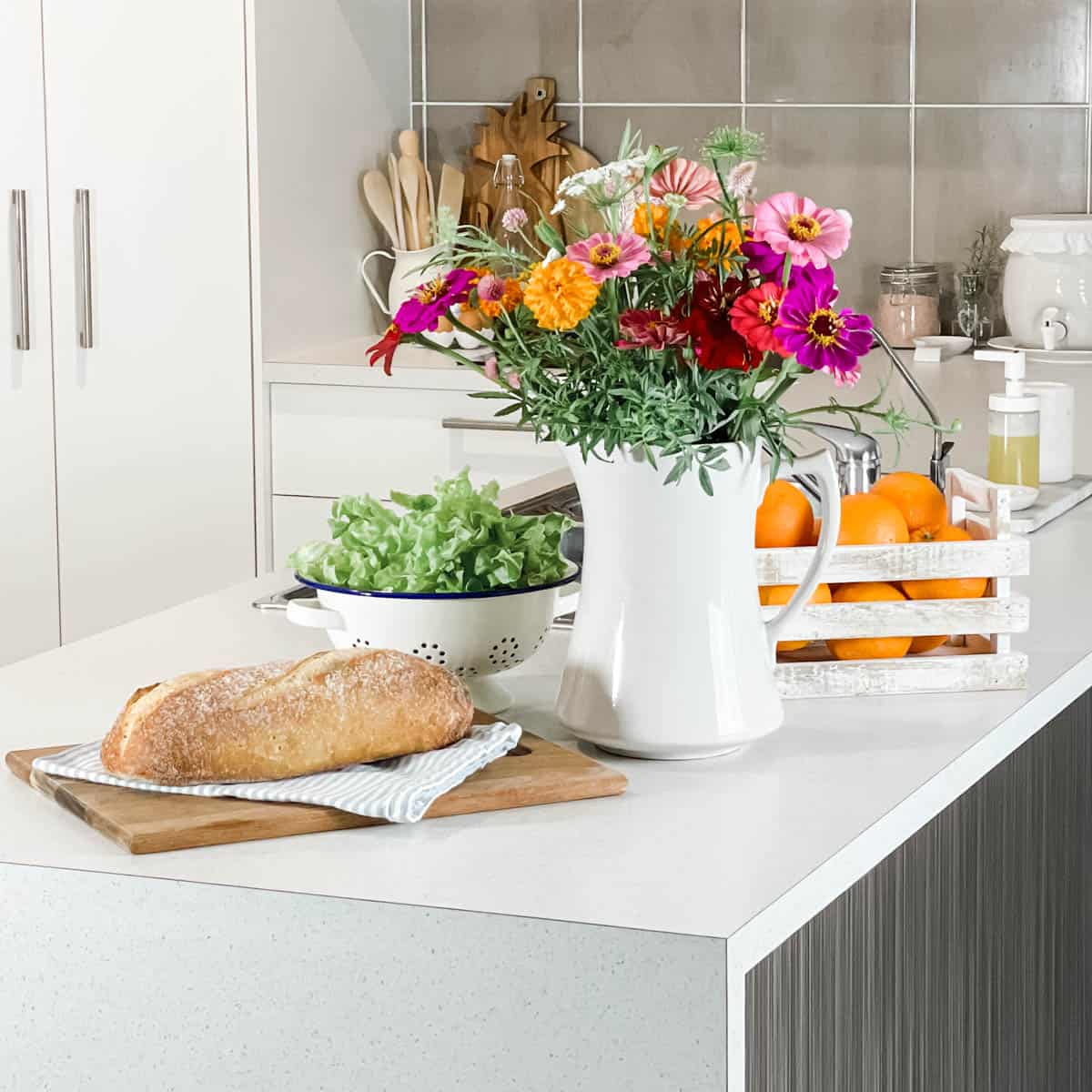 Picture of a jug of flowers, a loaf of bread and cutting board, tea bowl, colander with lettuce and a crate of oranges sitting on a kitchen bench