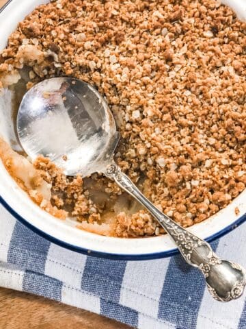 Close up image of a dish of apple crumble with a silver serving spoon set on a white and blue striped tea towel