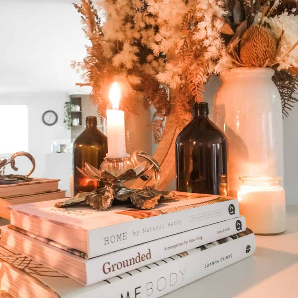 Arrangement of decorative items on a white mantle including a round mirror, stack of three white books with a candle on top, brown bottle, white vase with dried floral arrangement and glass candle