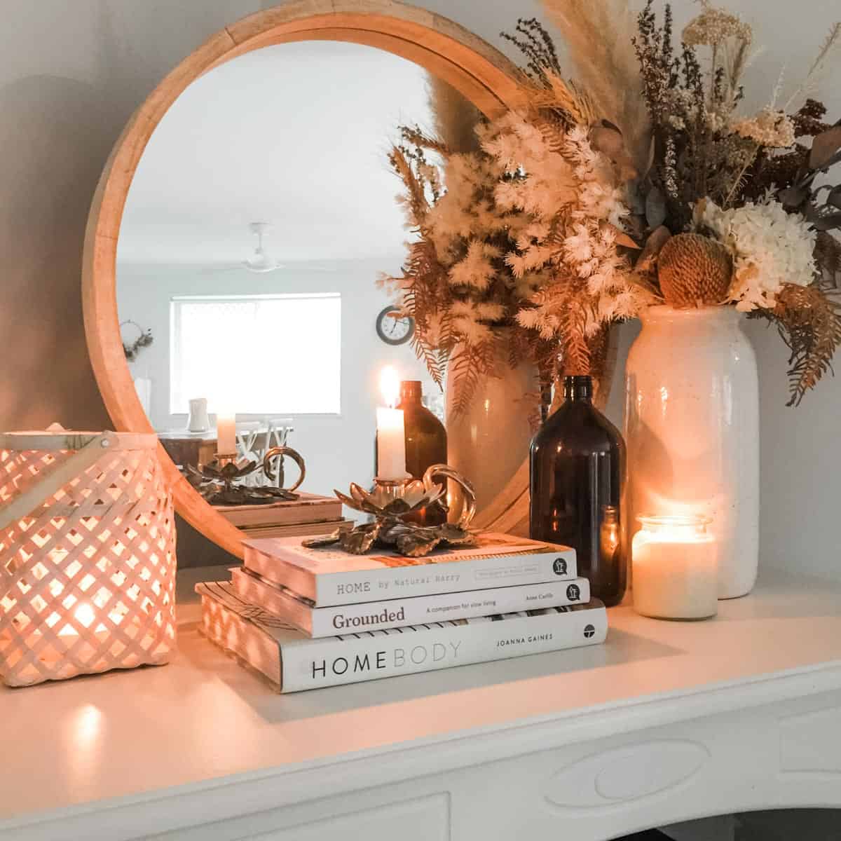 Arrangement of decorative items on a white mantle including a round mirror, stack of three white books with a candle on top, brown bottle, rattan lantern, white vase with dried floral arrangement and glass candle