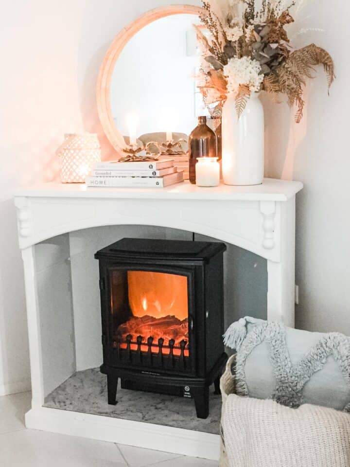 Image of a black faux fireplace hater in a white cabinet next to a basket with a cushion and throw rug and decorated with a round mirror, books, lantern candle and vase with dried florals