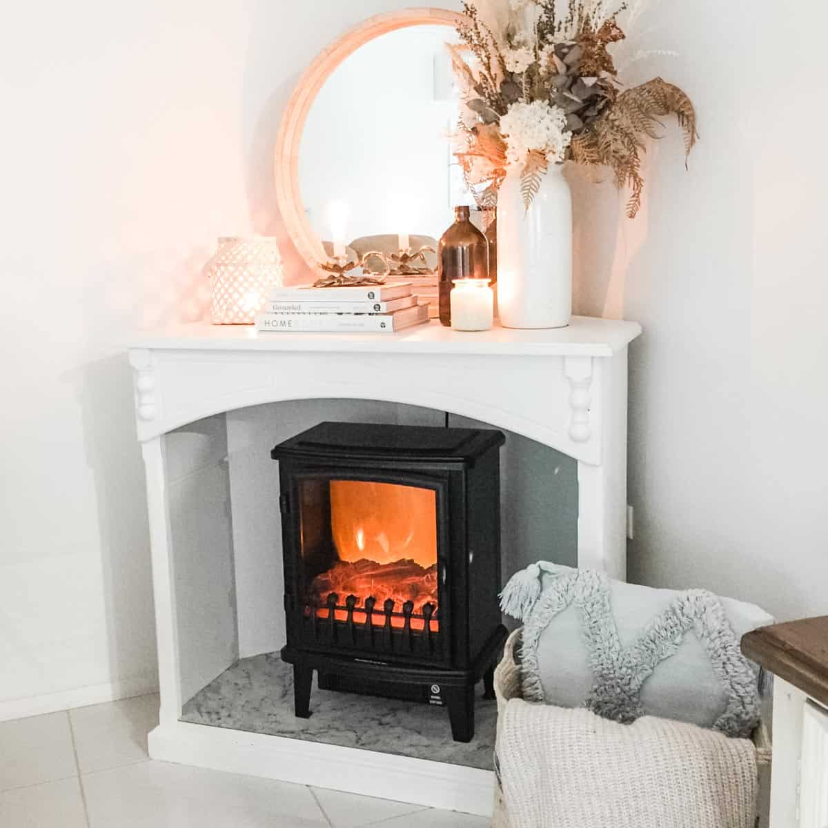 Image of a black faux fireplace hater in a white cabinet next to a basket with a cushion and throw rug and decorated with a round mirror, books, lantern candle and vase with dried florals