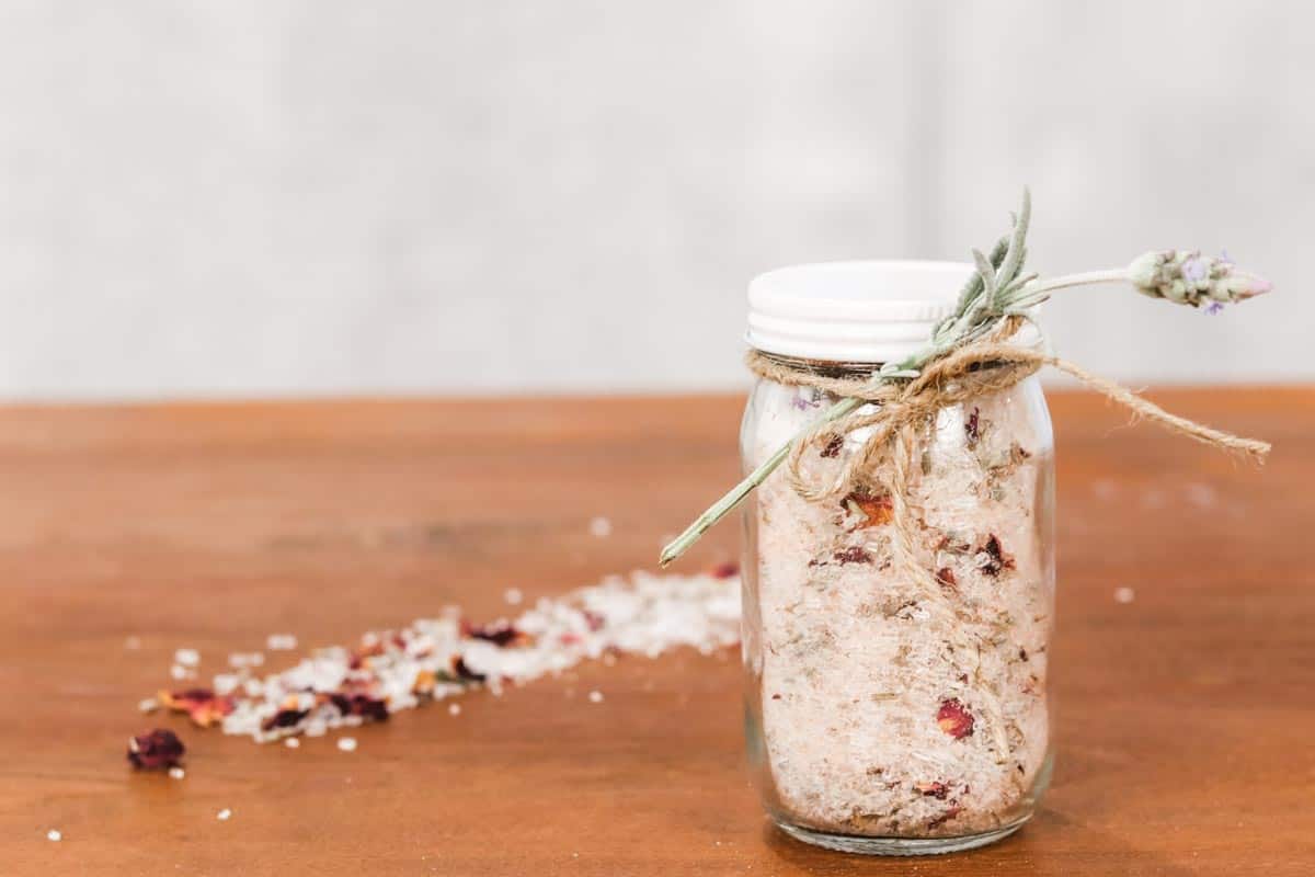 Image of a glass jar filled with bath salts, rose petals and lavender petals, tied with a twine bow and a sprig of lavender, on top of a timber table, next to a sprinkling of bath salts.