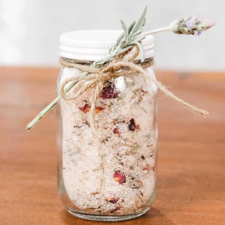 Glass jar with a white lid filled with white and pink salt crystals and dried rose petals and lavender petals. Tied with a bow of twine and adorned with a sprig of lavender.