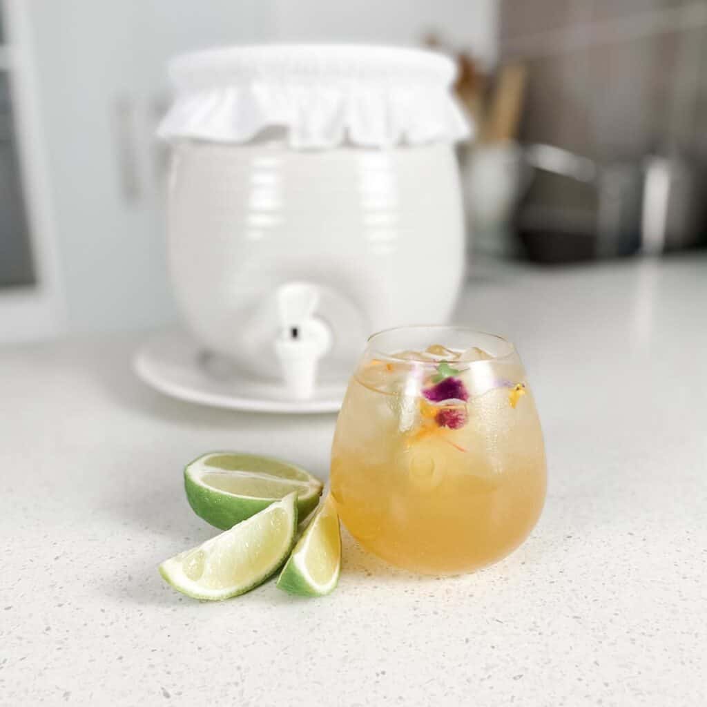 glass of kombucha next to slices of lime in front of a white stoneware fermentation crock brewing vessel