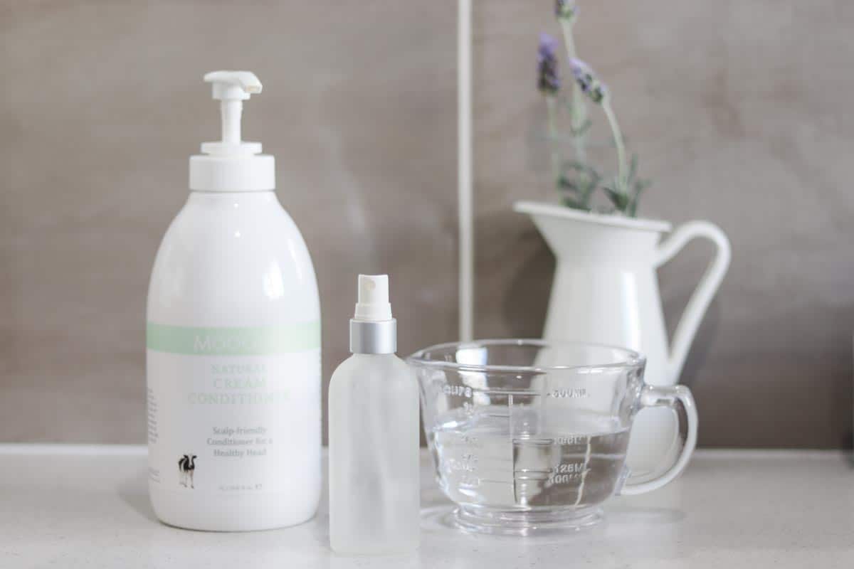 Image of pump bottle of hir conditioner next to an empty spray bottle and a jug of water. There is a white jug in the background with lavender