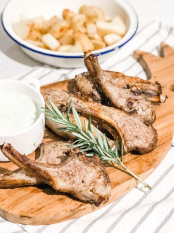 cooked lamb cutlets on a timber serving board with a grey and white striped teatoowl and a small white bowl of yogurt sauce and a bowl of roasted potato in the background