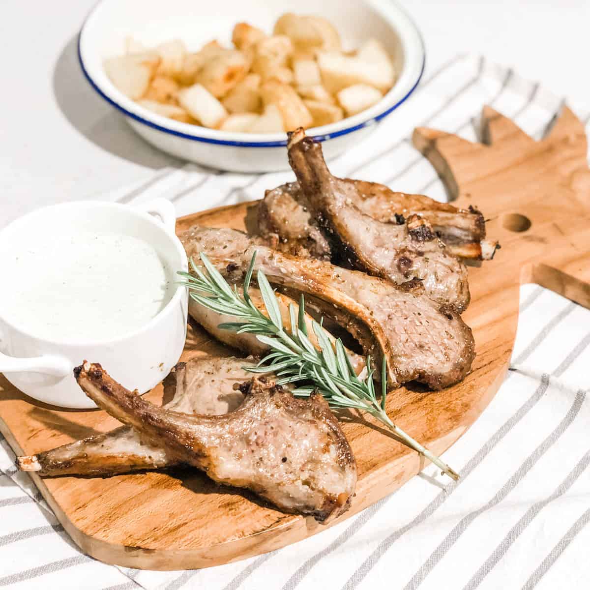 cooked lamb cutlets on a timber serving board with a grey and white striped teatoowl and a small white bowl of yogurt sauce and a bowl of roasted potato in the background