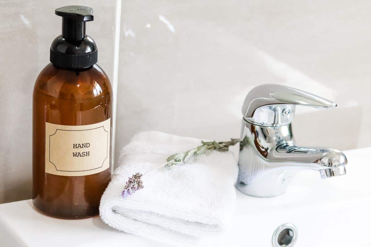 Amber foaming sopa dispenser on a white sink next to a white washcloth, sprig of lavender and silver tap