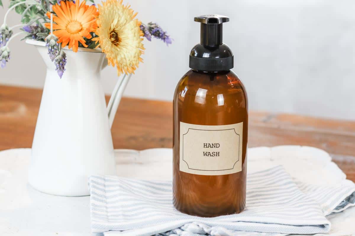 Amber coloured foaming hand soap pump dispenser with a label and text 'hand soap' on a white tray with a blue and white tea towel and a white pitcher of flowers