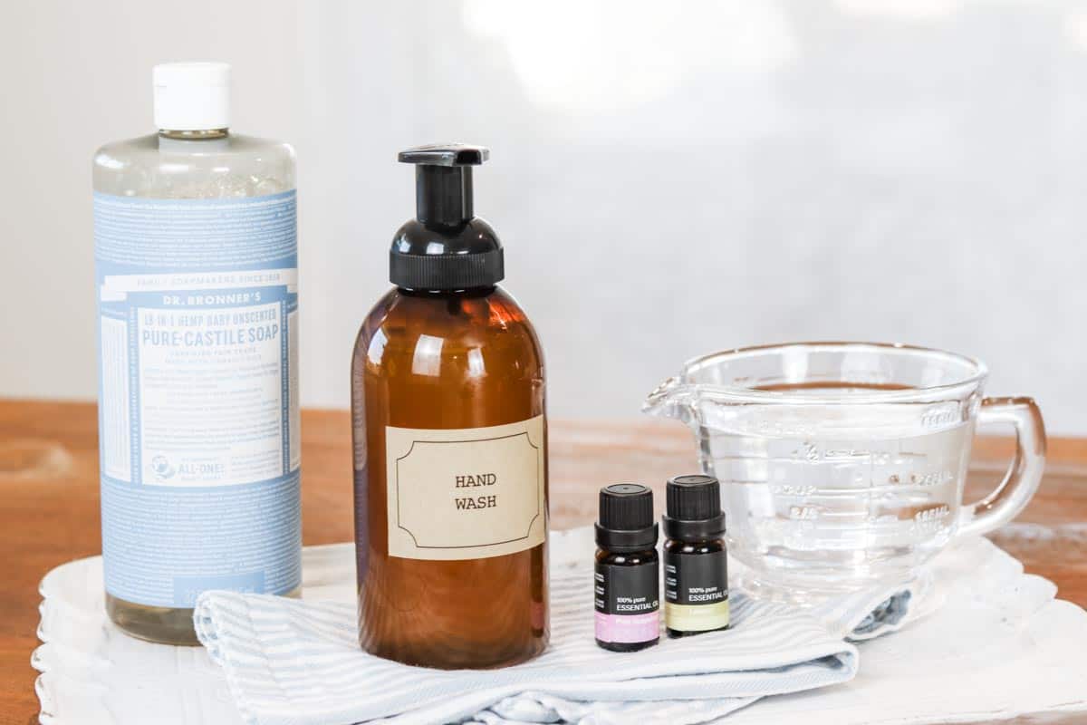 Group of ingredients including a bottle of Dr Bronner's Pure Castile soap, empty amber foaming soap dispenser, two bottles of essential oils and a jug of water on a blue and white striped tea towel and white tray