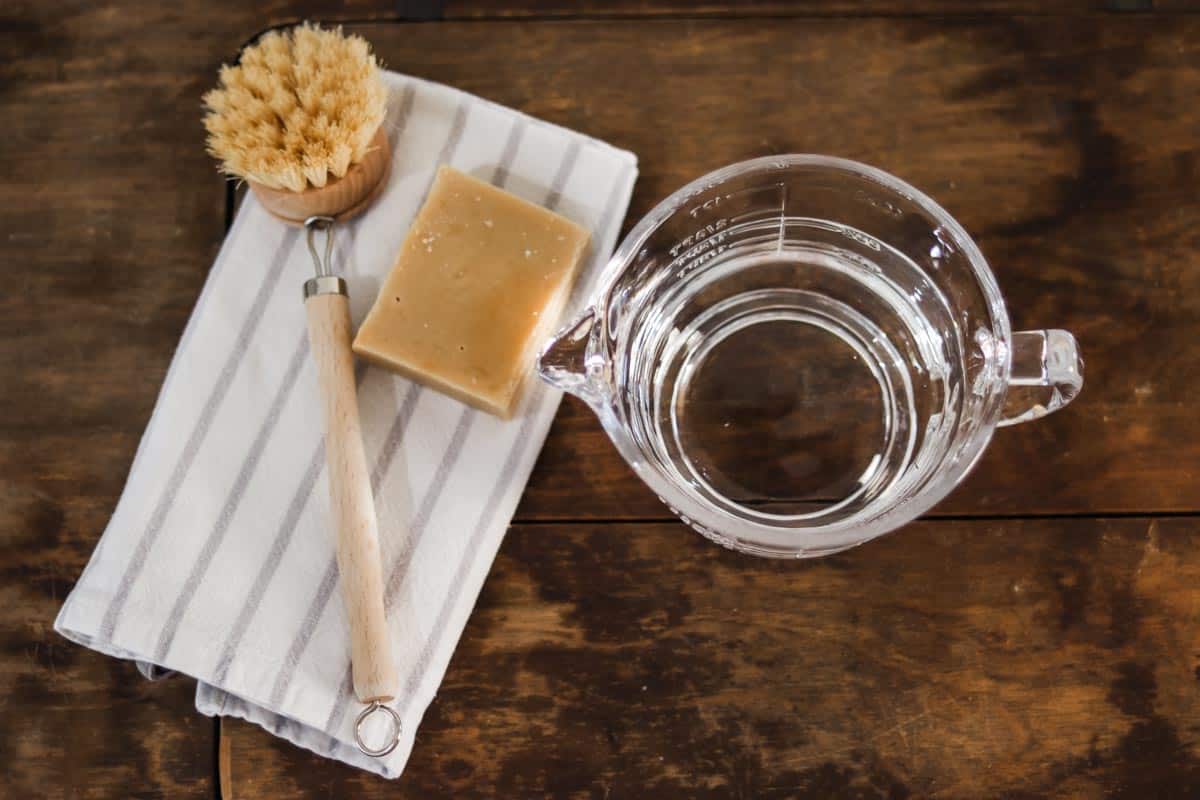 Overhead image of a glass jug full of water next to a white and grey striped tea towel, scrubbing brush and a bar of soap