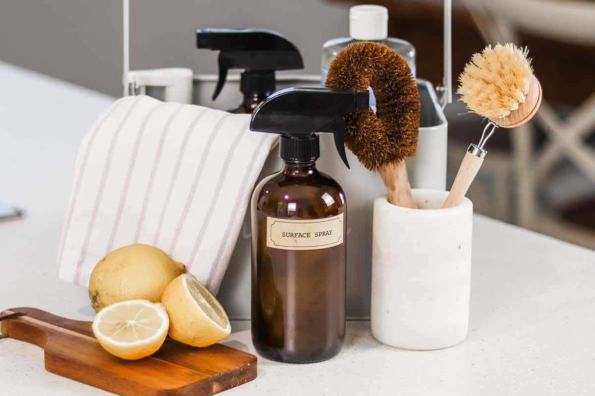Image of a group of natural cleaning materials including brushes, spray bottles and a halved lemon on a bench in front of a caddy