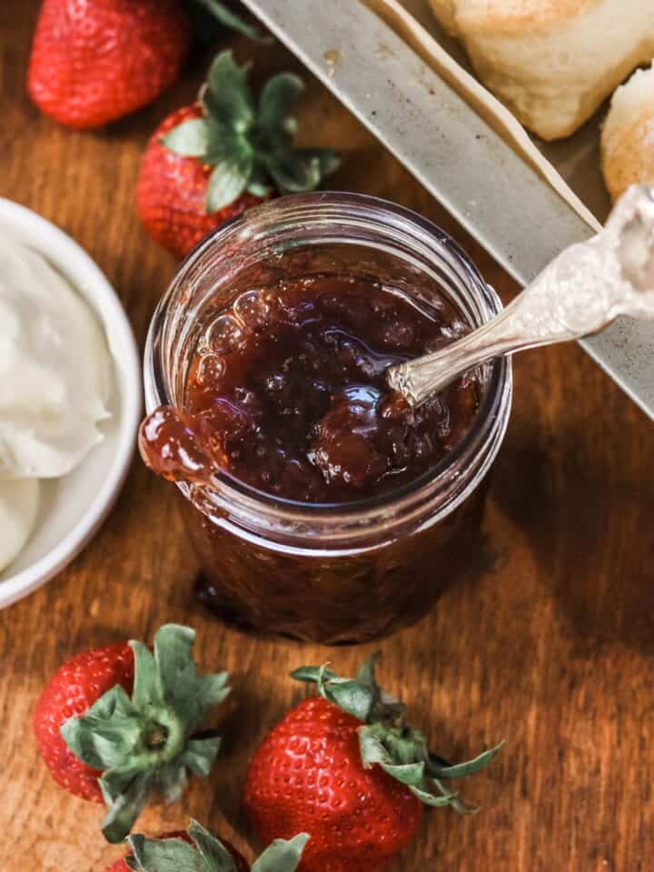 Overhead image of an open jar of strawberry jam next to a dish of cream and a tray of scones