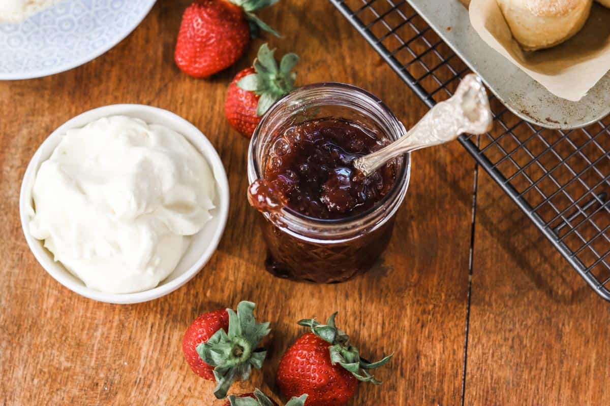 Overhead image of an open jar of strawberry jam next to a dish of cream and a tray of scones