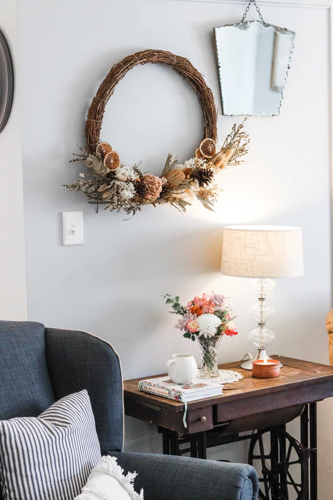 Image of neat and tidy living room with armchair, cushions, next to a table with a book, cup of tea, vase of flowers, lamp and candle. There is a mirror and wreath hanging on the wall