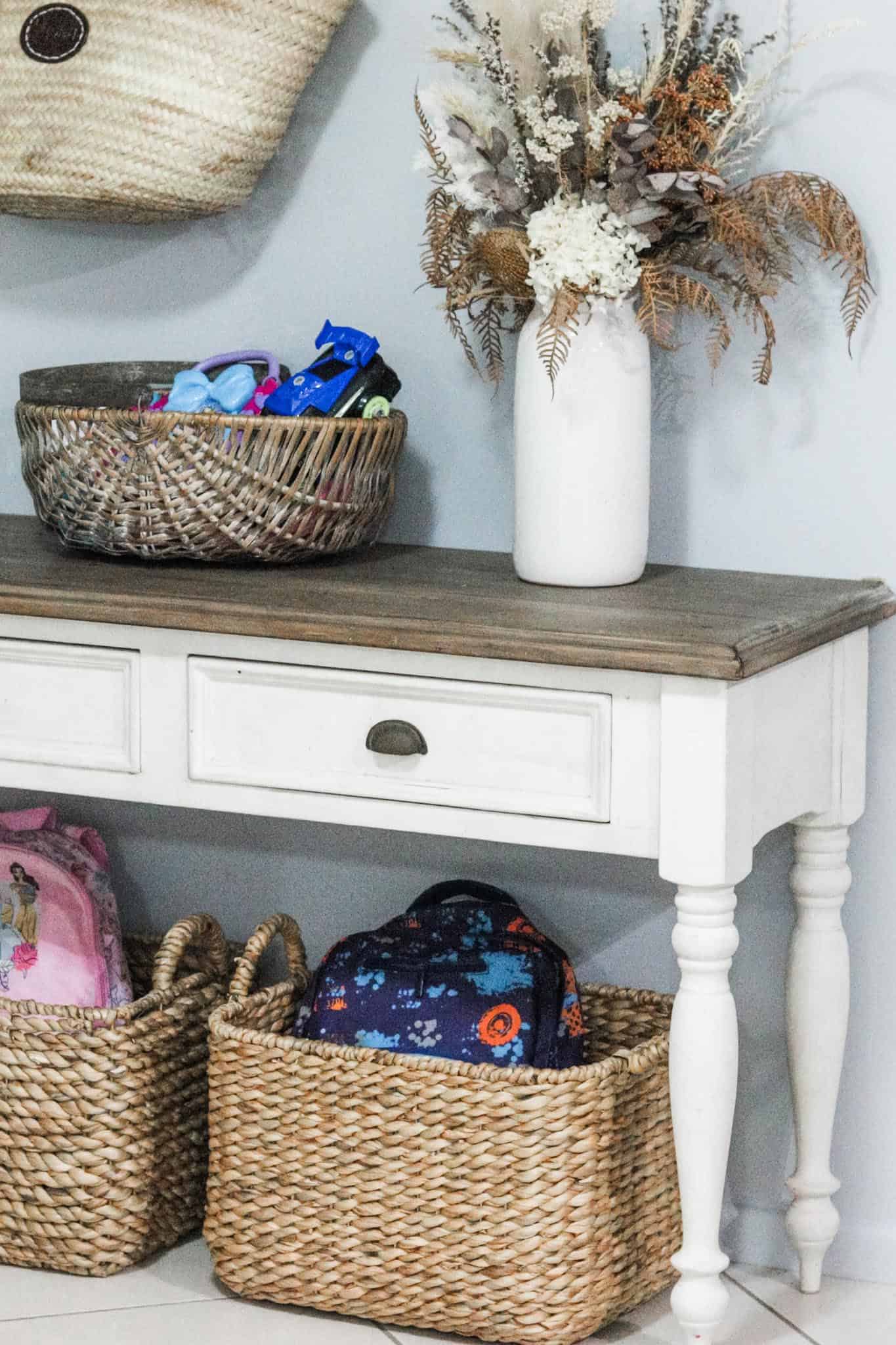 Image of a console table with two large seagrass baskets underneath. The baskets are holding school bags. On top of the table is a basket full of toys and a vase with a dried floral arrangement. There is a straw basket hanging above the table.