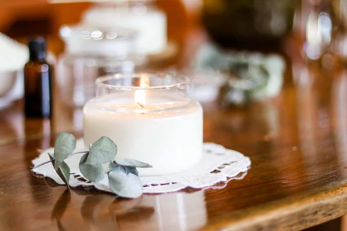 Close up image of a glass soy wax container candle which is lit, sitting on a cotton and lace doily, next to eucalyptus leaves with candle making supplies in the background