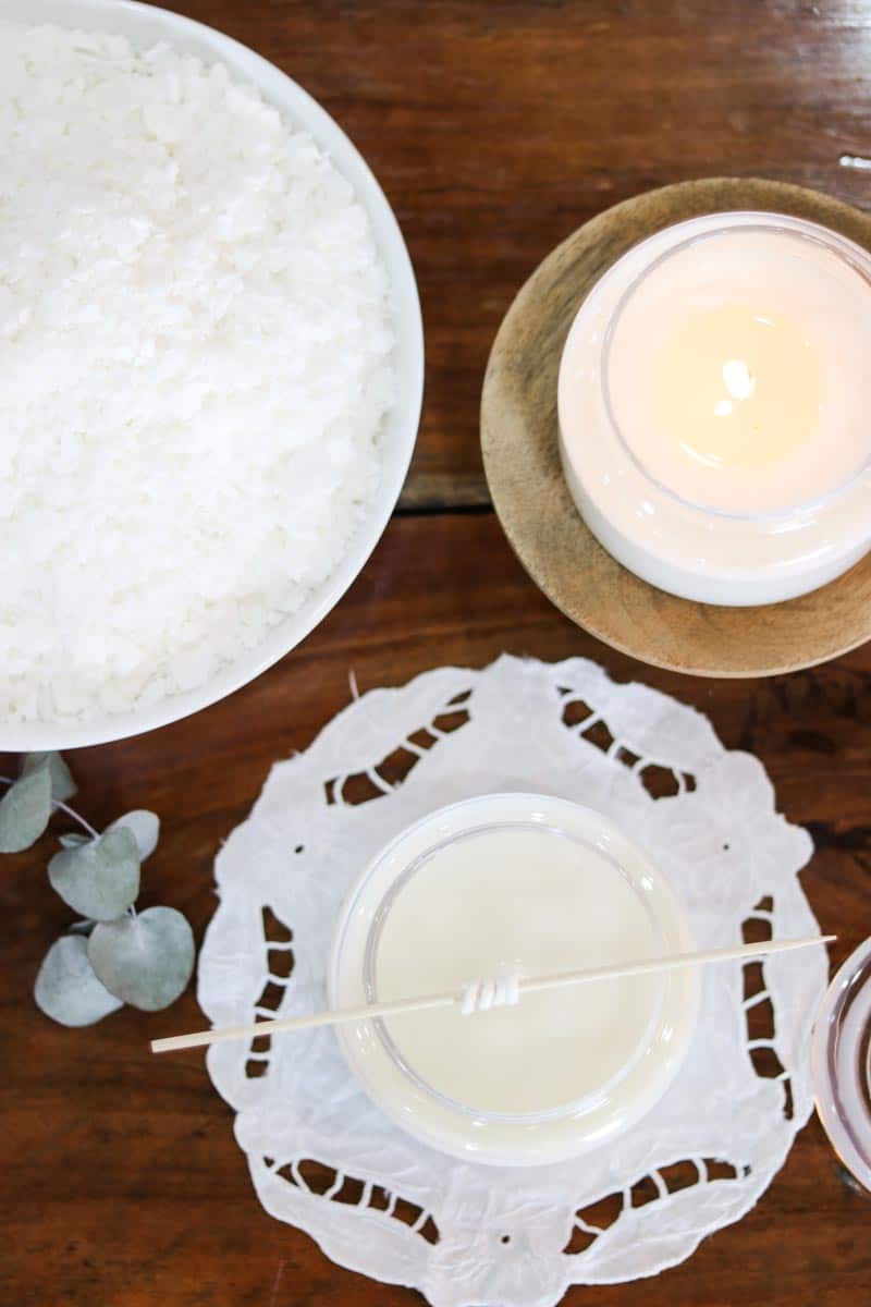 Overhead image of a glass soy wax container candle which is lit, sitting on a wooden dish, next to another candle sitting on a cotton and lace doily with its wick still wound around a skewer, next to eucalyptus leaves and a bowl of soy wax flakes
