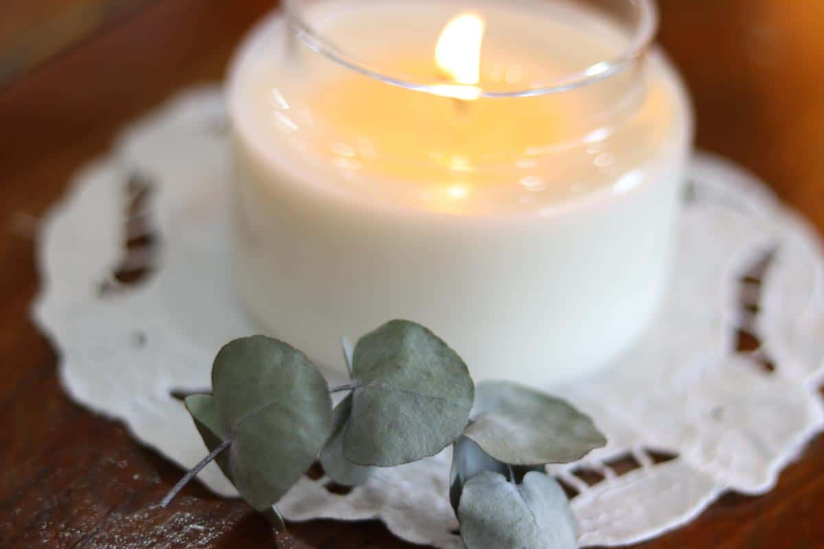 Close up image of a glass soy wax container candle which is lit, sitting on a cotton and lace doily, next to eucalyptus leaves