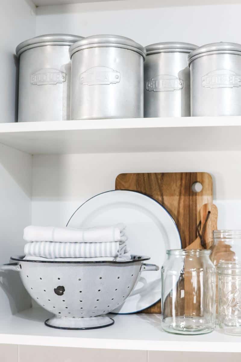 Image of two shelves, one filled with vintage aluminium canisters and the other with a vintage colander filled with folded blue and white tea towels, a blue and white enamel plate, two wooden serving boards and a selection of empty glass jars