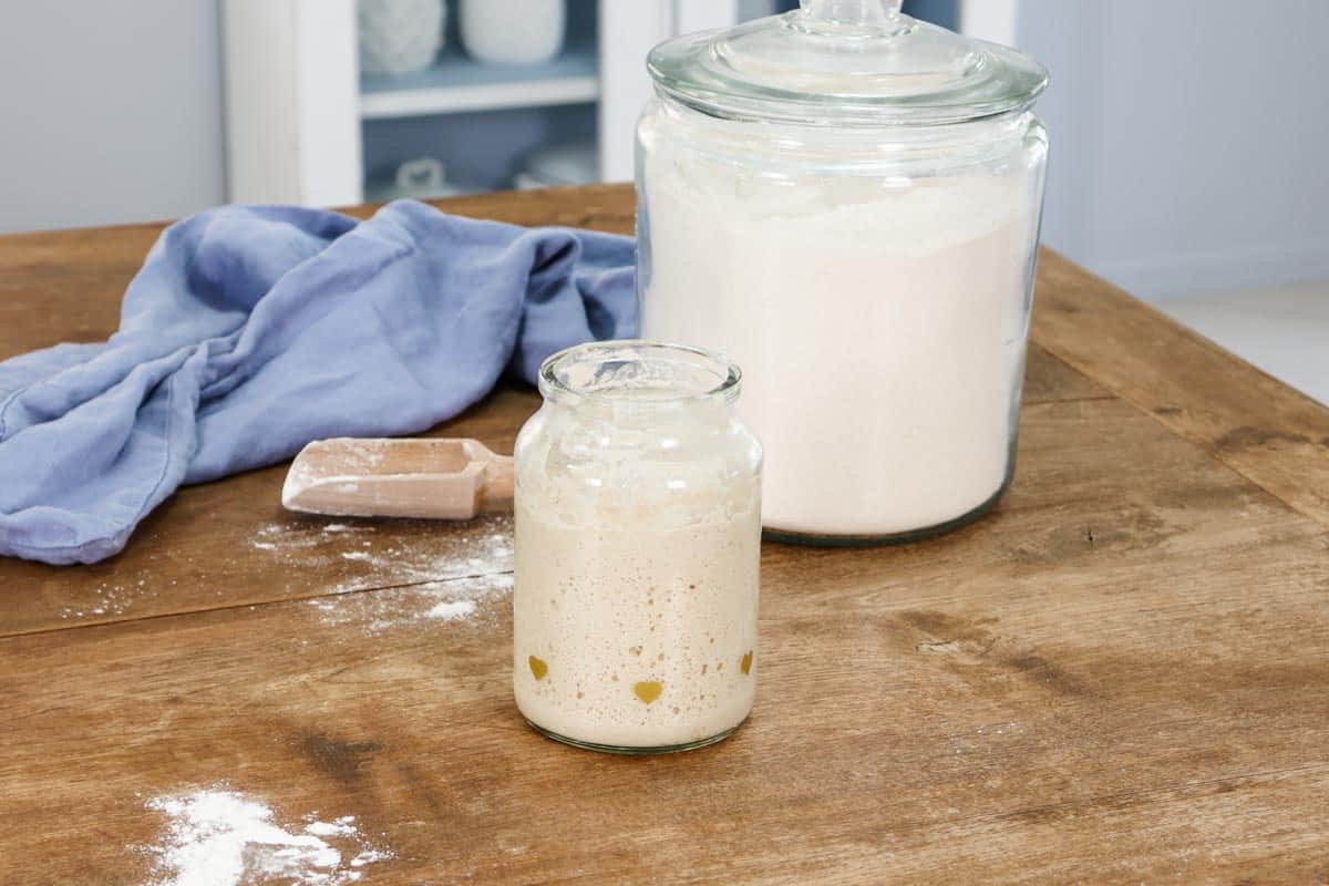 Image of a glass jar of sourdough starter next to a flour scoop and a large canister of flour