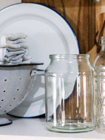 Image of a group of empty glass jars, an enamel colander filled with blue and white folded tea towels, a white and blue enamel plate, two wooden serving boards arranged on a shelf