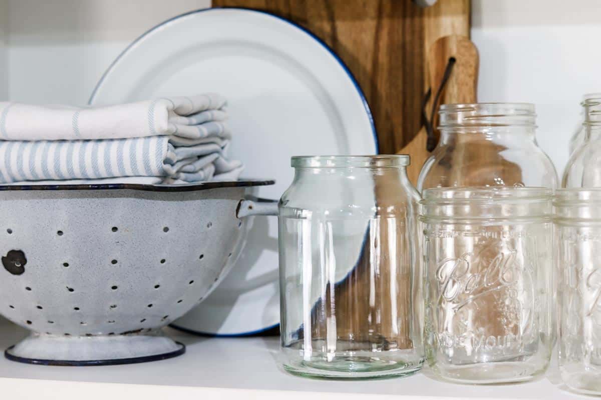 Image of a group of empty glass jars, an enamel colander filled with blue and white folded tea towels, a white and blue enamel plate, two wooden serving boards arranged on a shelf