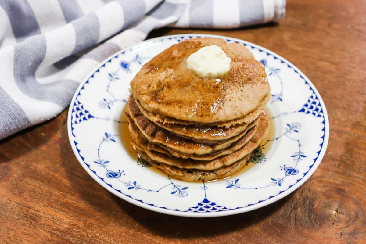 Image of a stack of sourdough pancakes on a blue and white plate topped with butter and maple syrup set on top of a wooden table with a blue and white tea towel