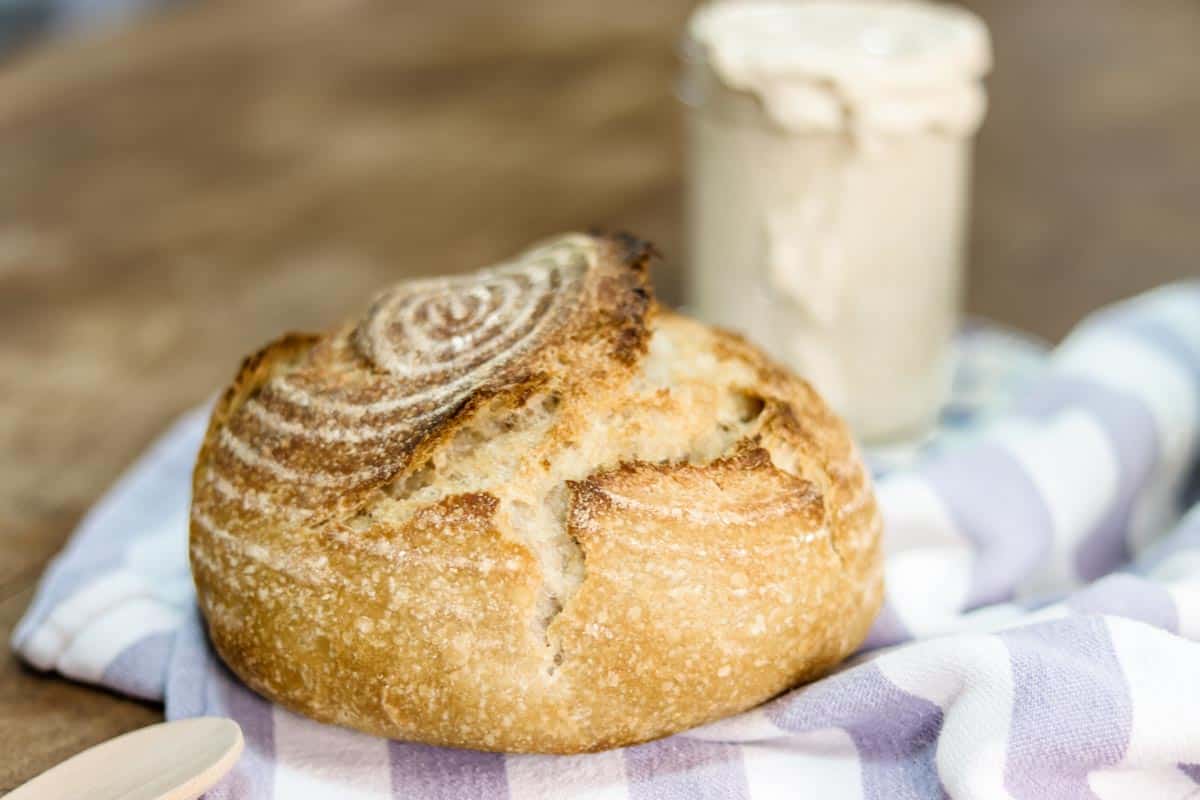 Freshly baked loaf of sourdough bread on a blue and white cloth in the foreground, and a jar of sourdough starter in the background