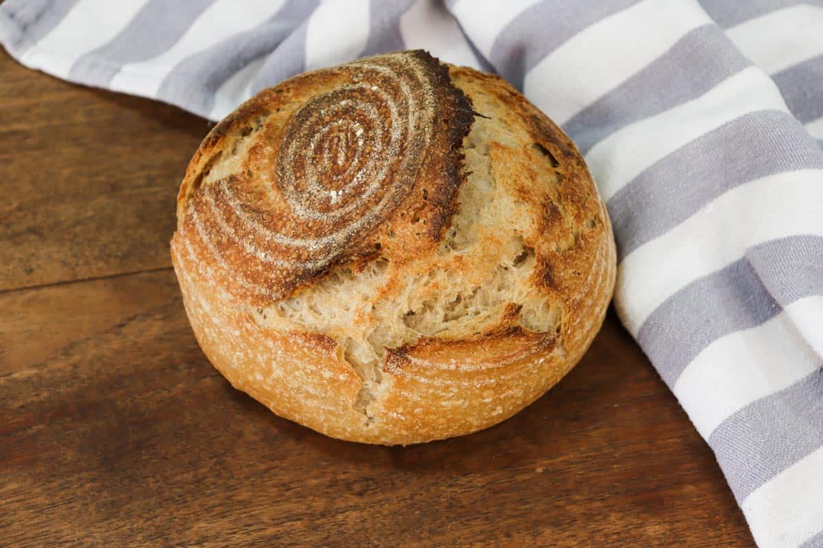 A loaf of sourdough bread sitting on a wooden table next to a white and blue tea towel showing what makes sourdough bread looks different