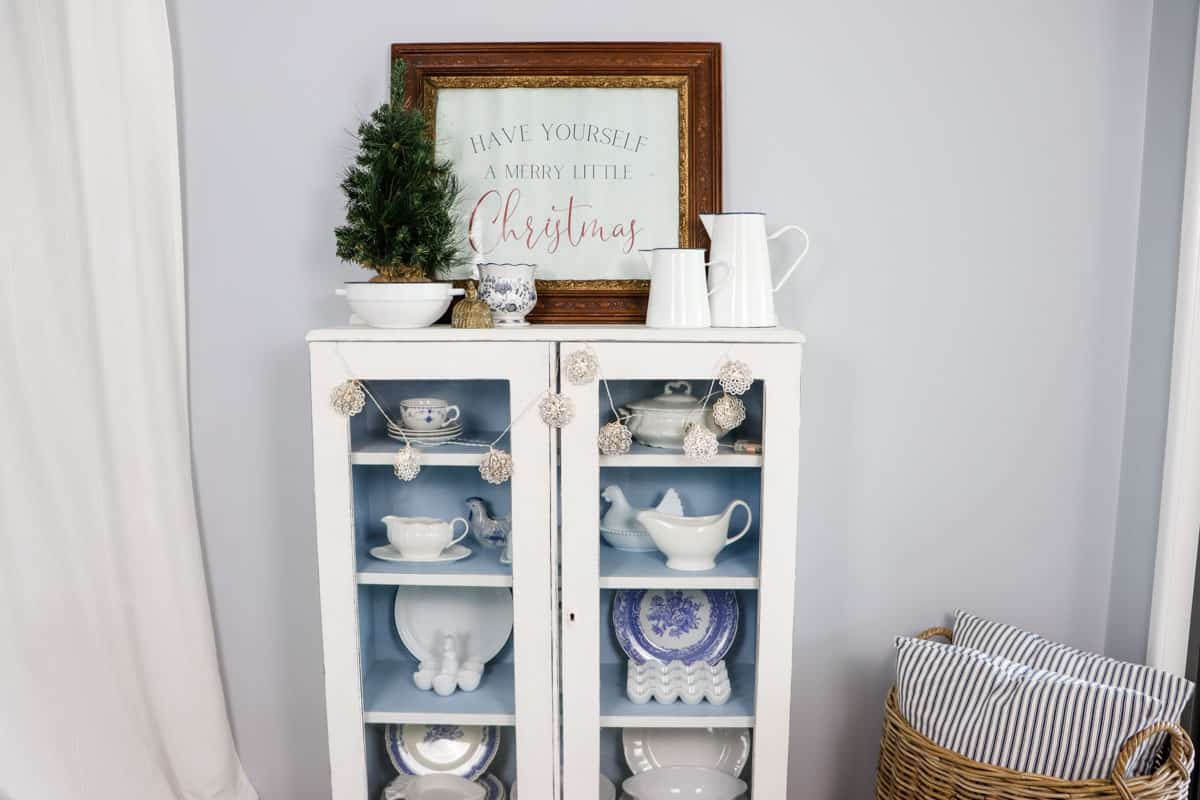 The china hutch is decorate with a vintage frame with poster print, a small Christmas tree in a thrifted enamel bowl, a blue and white vase, brass bell, two enamel pitchers and lights