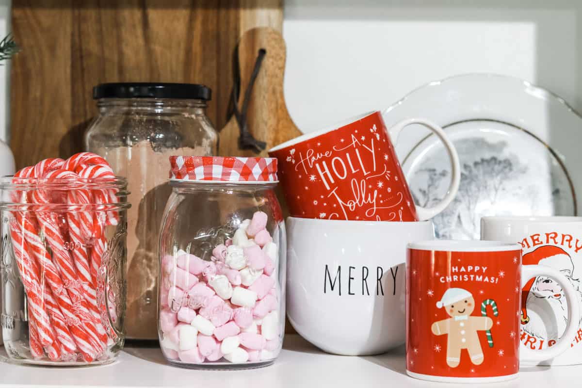 a collection of jars filled with hot cocoa powder, marshmallows, and candy canes next to an assortment of Christmas themed mugs