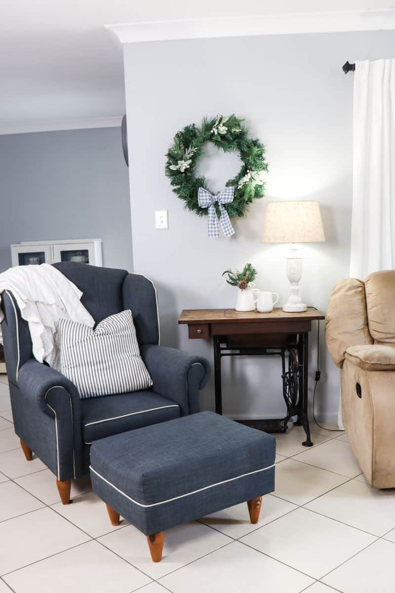 A blue reading chair with cushion and crochet throw rug next to a lamp table with a lamp, thrifted pitcher, and DIY cottage style Christmas wreath