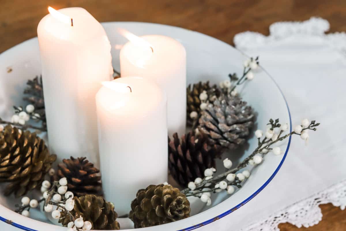 A close up image of a cottage style table centrepiece with candles and pinecones set on a thrifted white crochet edge table runner