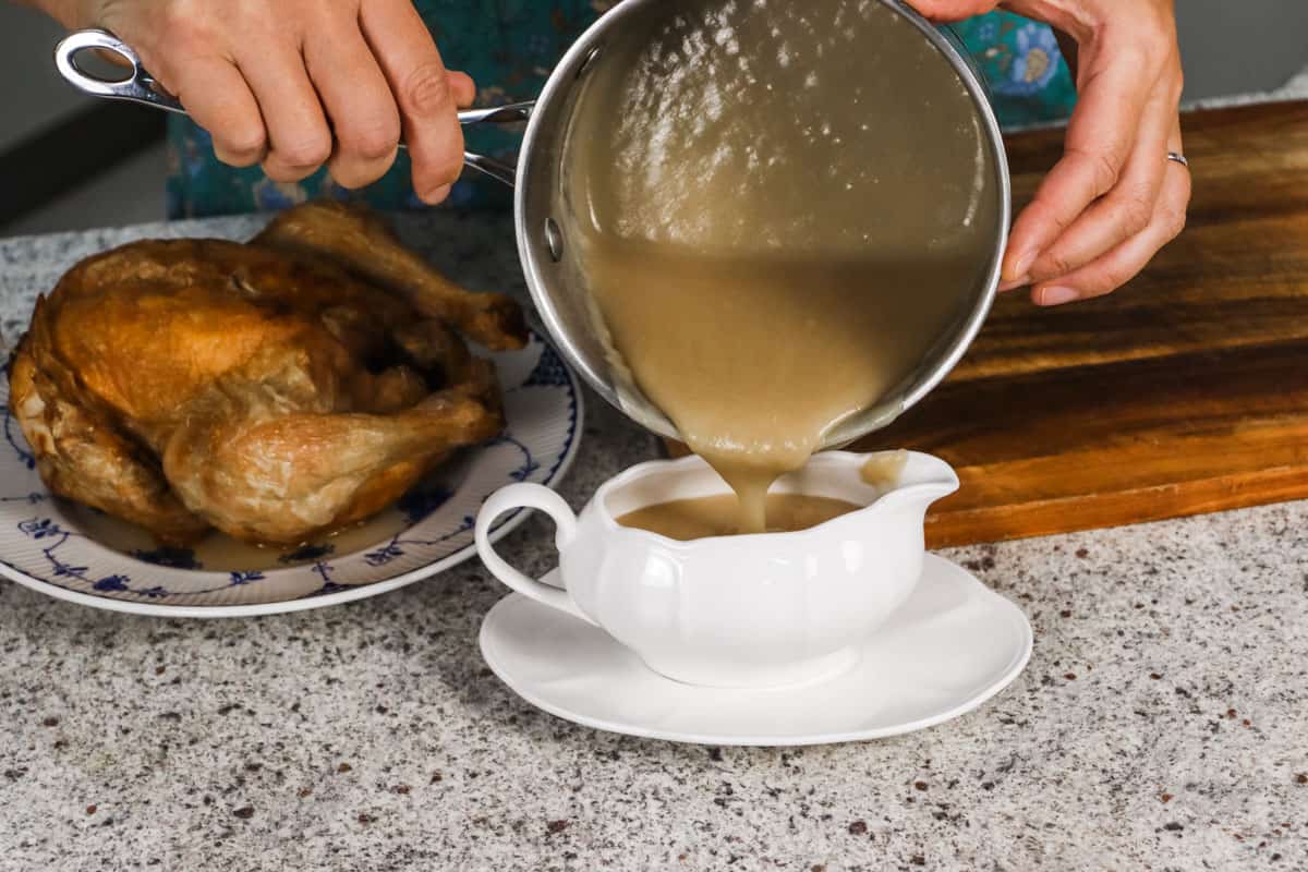 the gravy in the saucepan is being poured into a jug, next to a plate with a roasted chicken