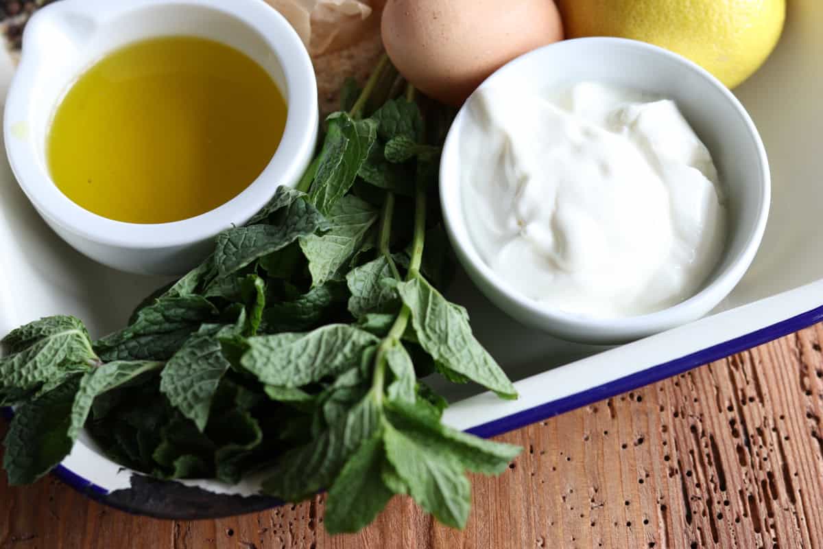A small bowl of yogurt sits on an enamel tray next to other ingredients to make crumbed lamb cutlets in the air fryer including extra virgin olive oil, fresh mint, a egg and lemon