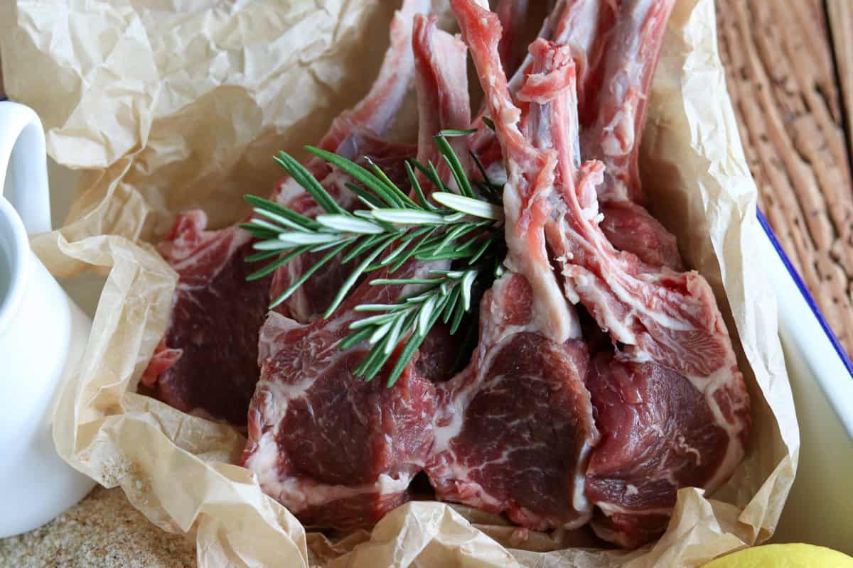 Raw lamb cutlets have been trimmed/frenched and are sitting on a piece of parchment paper garnished with fresh rosemary
