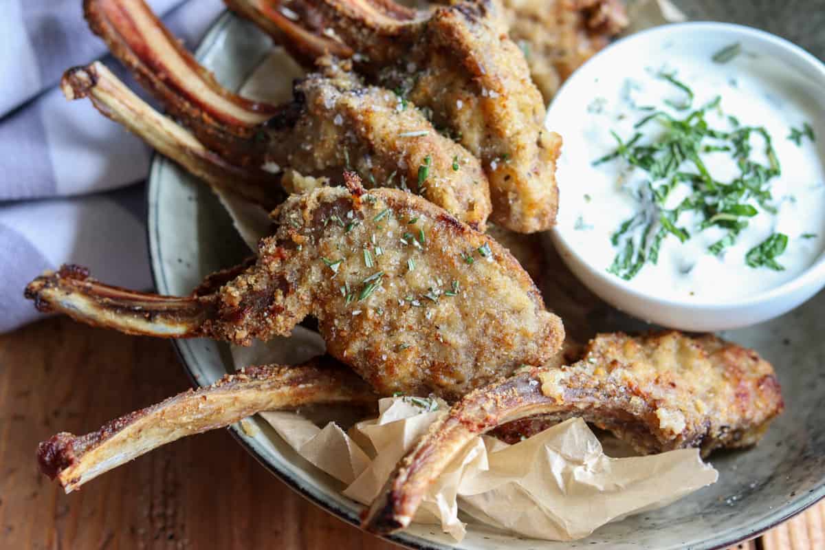 Crumbed lamb cutlets that have been cooked in the air fryer and garnished with fresh rosemary and salt flakes rest on a dish next to a small bowl of yogurt and mint sauce