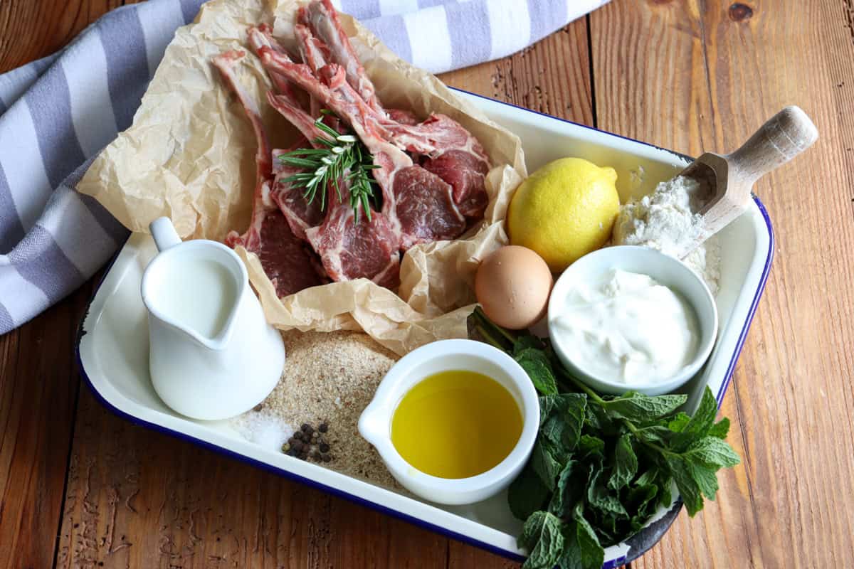 Ingredients to make crumbed lamb cutlets sit on an enamel tray including raw lamb cutlets, milk, salt, pepper, extra virgin olive oil, breadcrumbs, mint, rosemary, egg, yogurt, lemon and flour