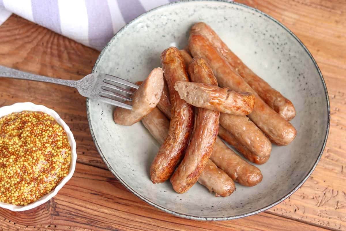Sausages that have been cooked in the air fryer sit in a bowl with a fork next to a bowl of mustard