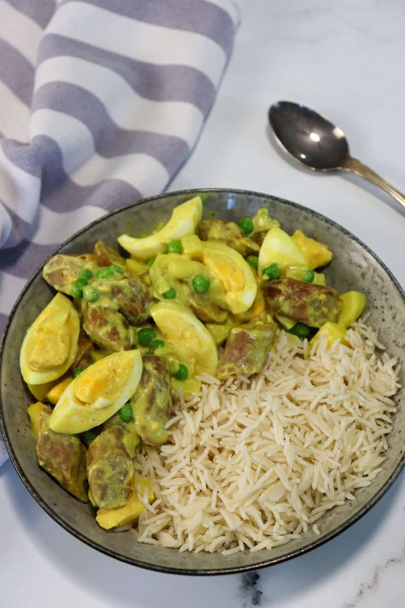 Old fashioned curried eggs and sausages plated in a dish with rice, set on a table with a tea towel and spoon