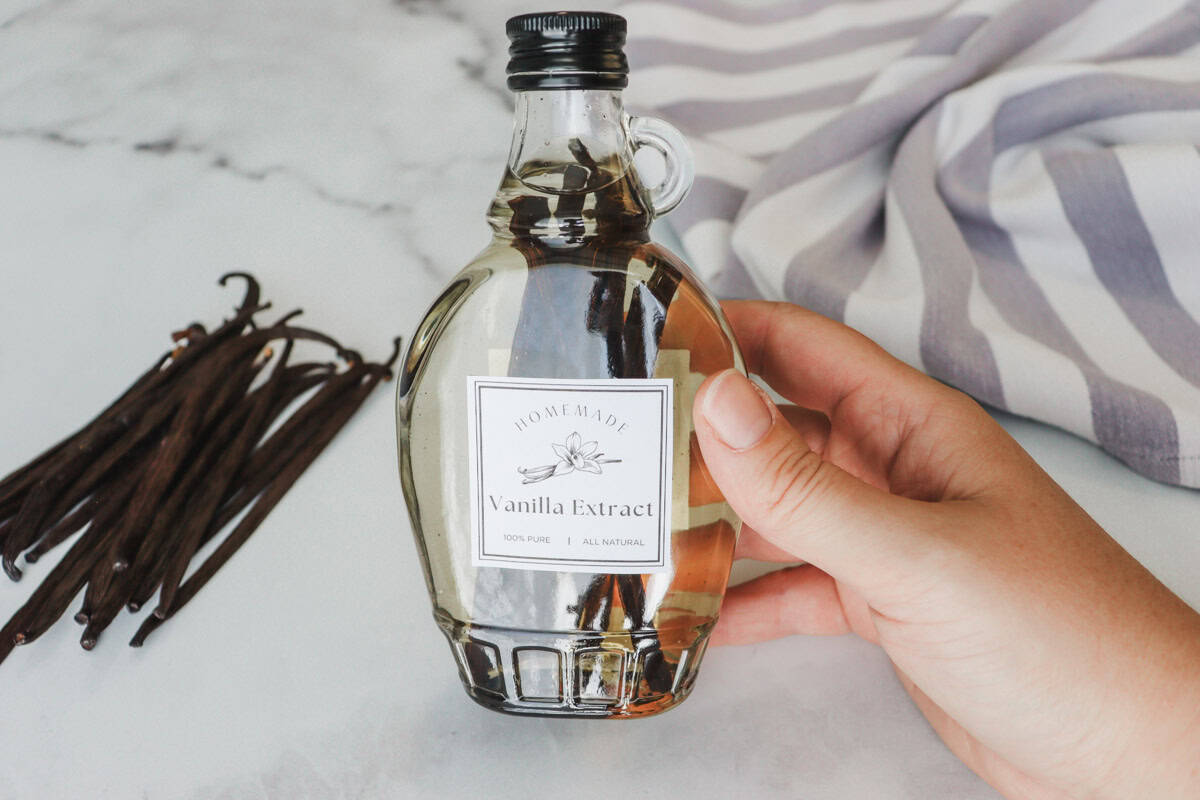 A hand holds a bottle of freshly made vanilla extract with a marble table top, dried vanilla beans and a tea towel in the background