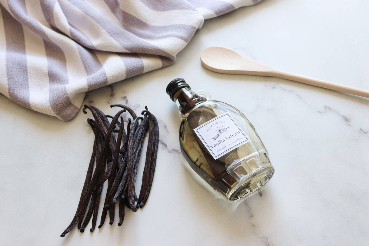 A bottle of freshly made pure homemade vanilla extract on a marble surface set next to a handful of dried vanilla beans, tea towel and a wooden spoon