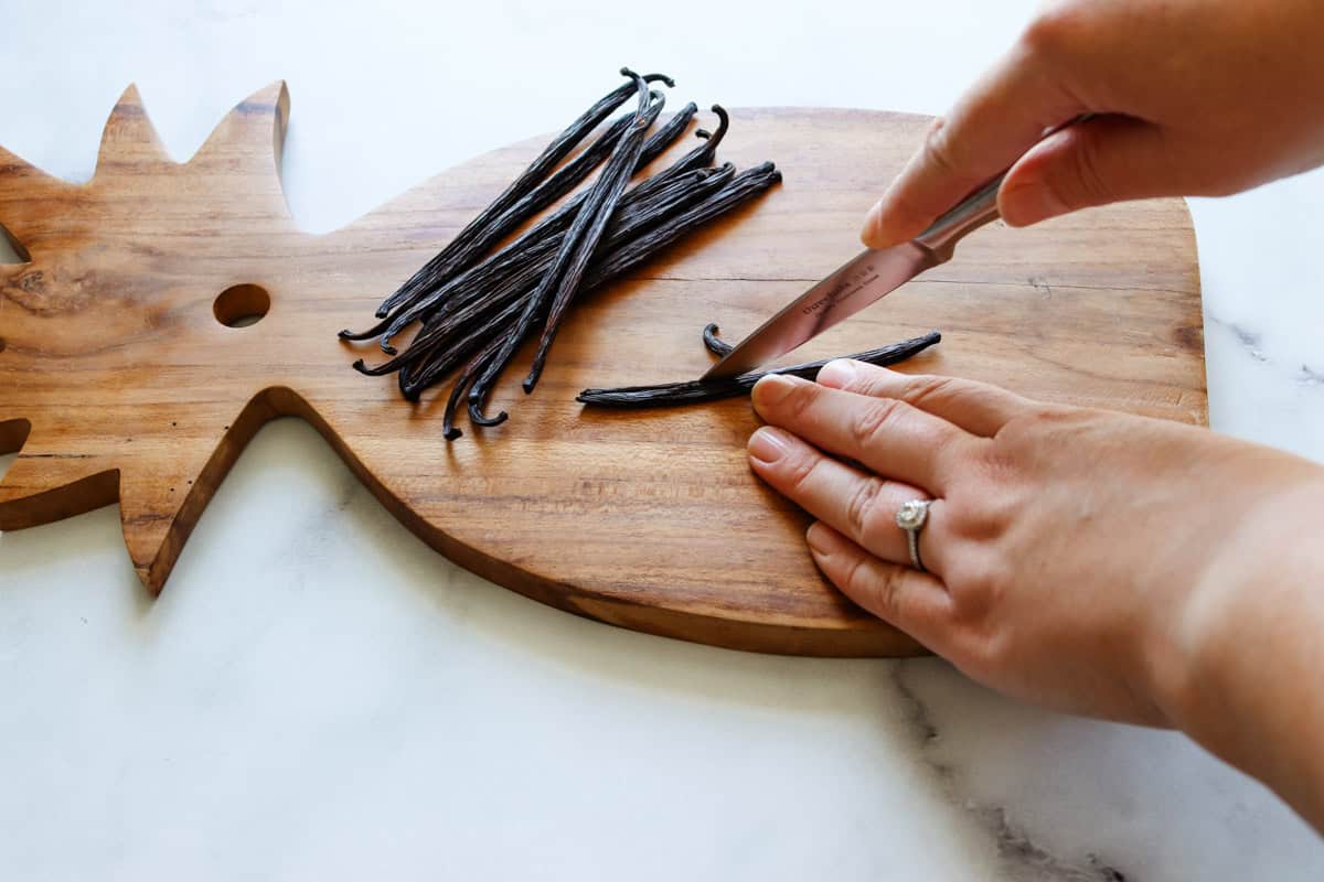 Dried vanilla beans are sliced lengthwise on a wooden chopping board using a paring knife