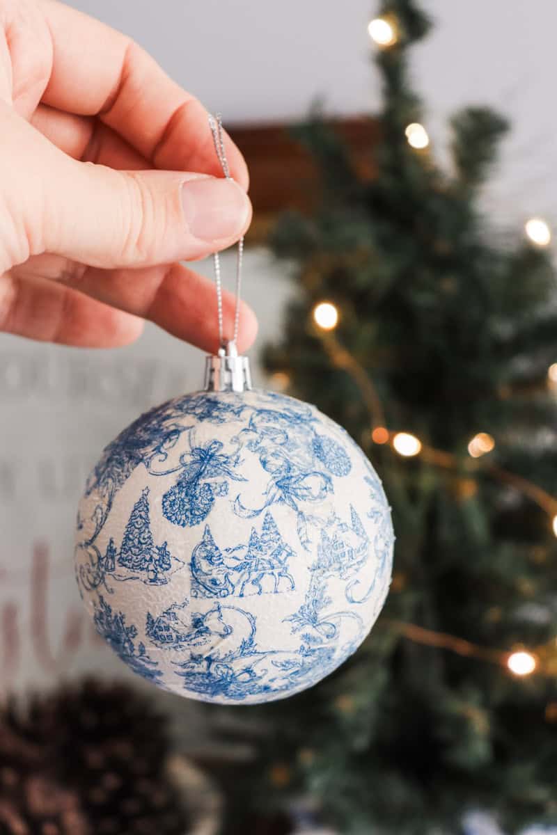Close up image of DIY blue and white paper napkin decoupage Christmas ornament being held in front of a Christmas tree