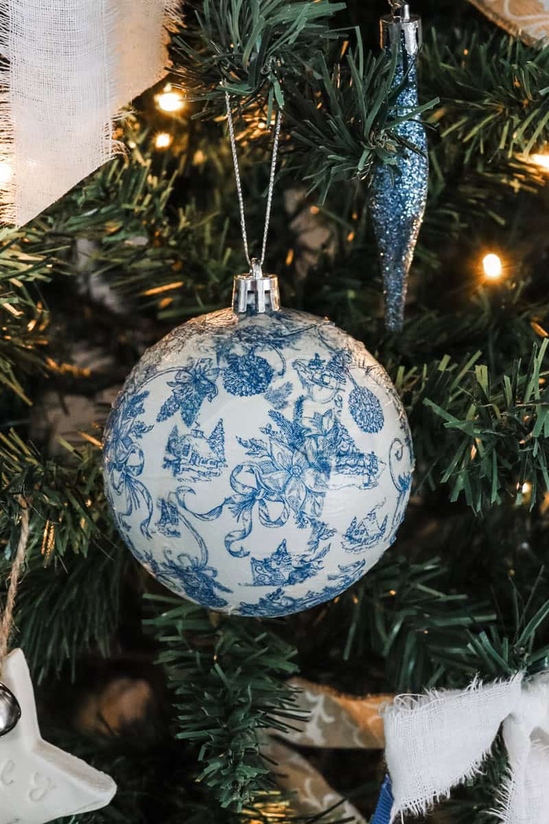 A decoupage Christmas ornament hangs on the Christmas tree surrounded by fairy lights and other ornaments in blue and white