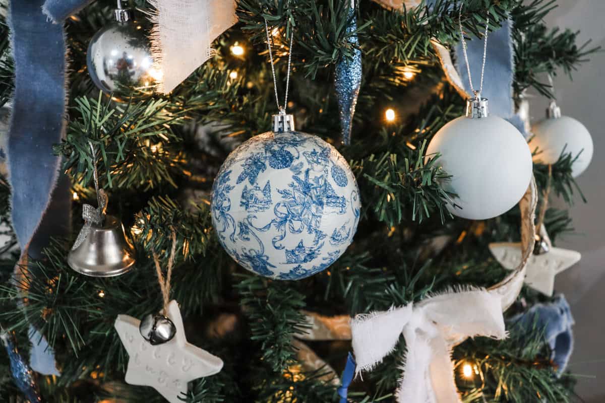 A decoupage Christmas ornament hangs on the Christmas tree surrounded by fairy lights and other ornaments in blue and white