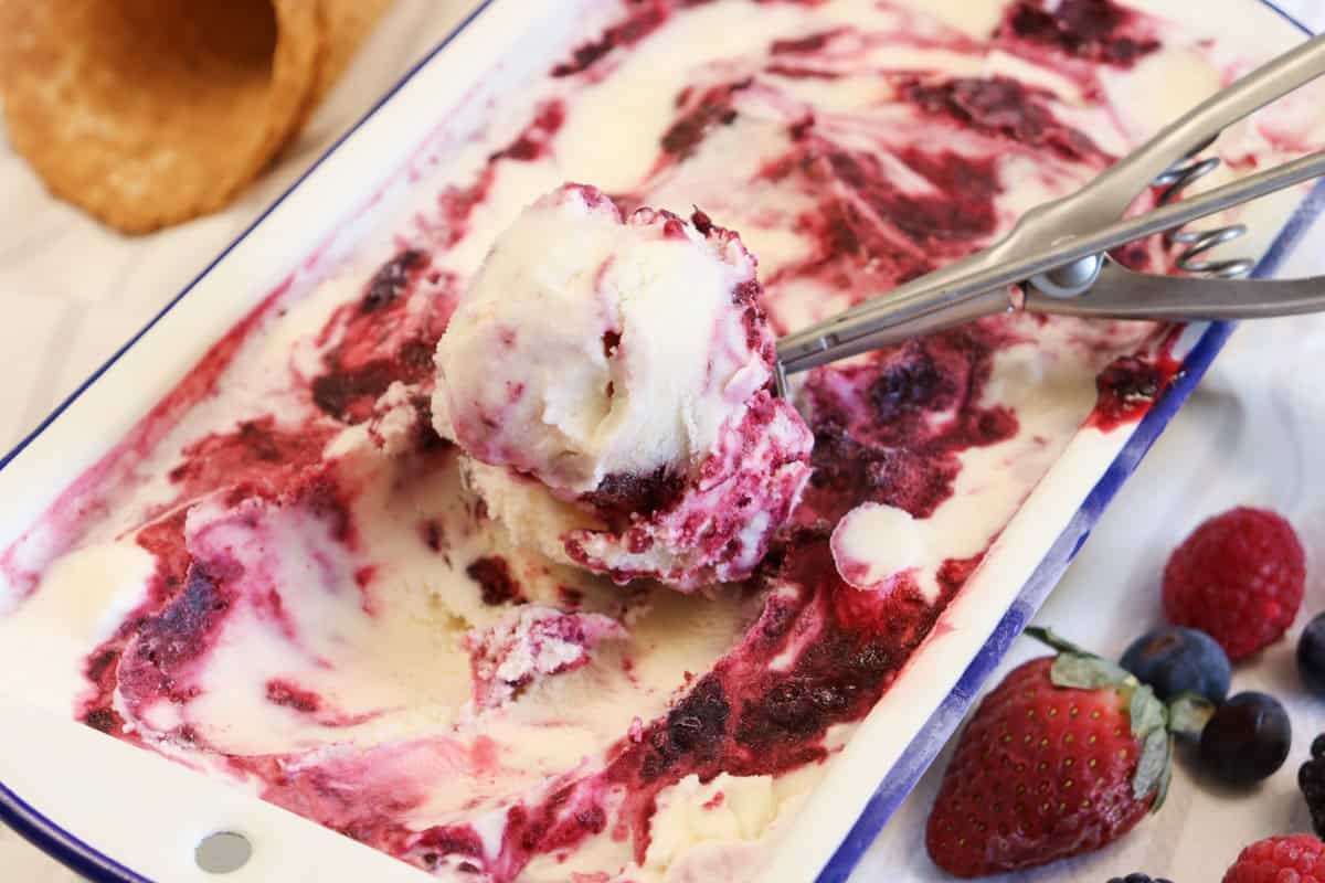 homemade berry ice cream being scooped out of the container surrounded by berries and waffle cones